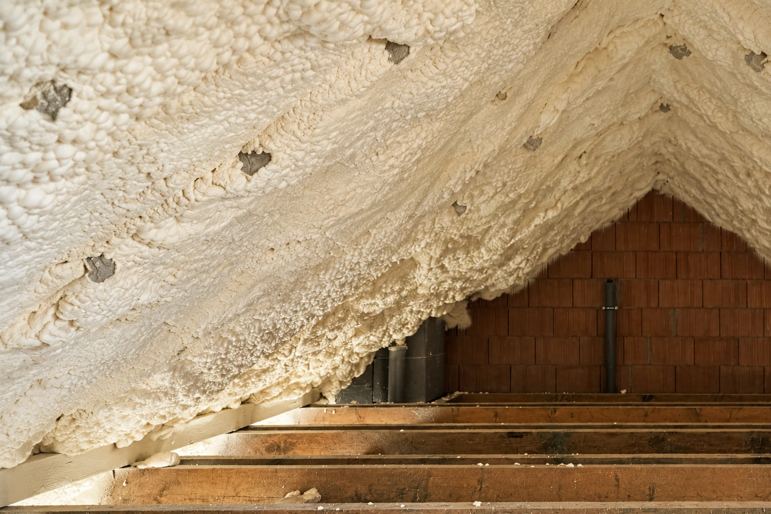 Thick white spray foam coats a sloped attic roof above exposed floor joists and a brick gable wall.