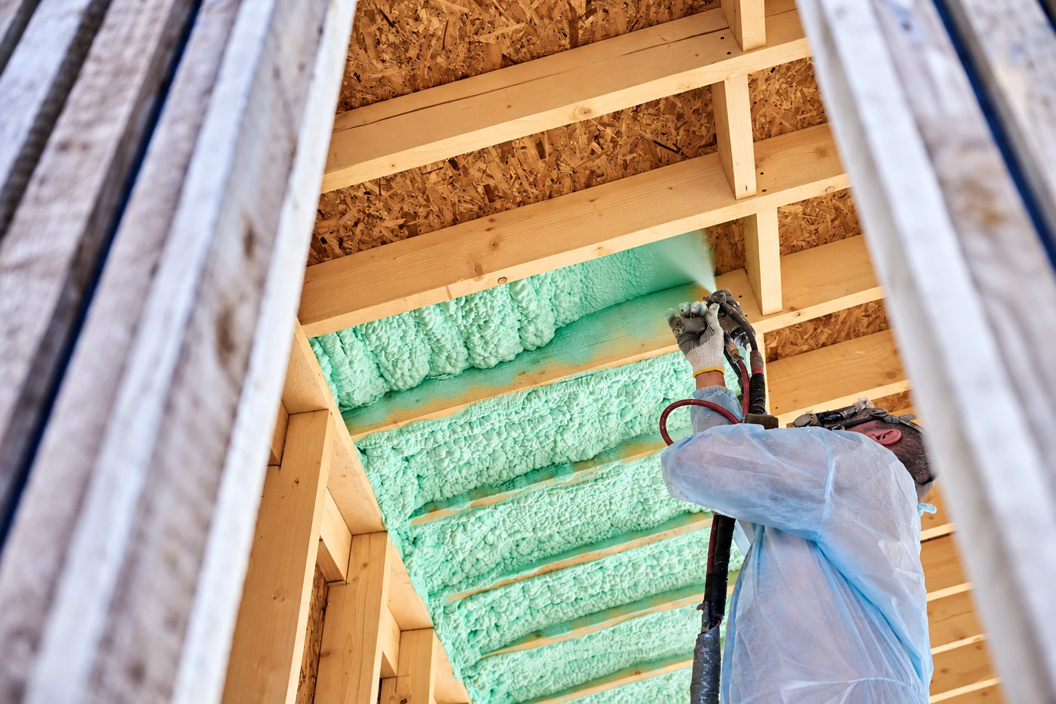 A worker in protective gear sprays green foam insulation between exposed ceiling joists.