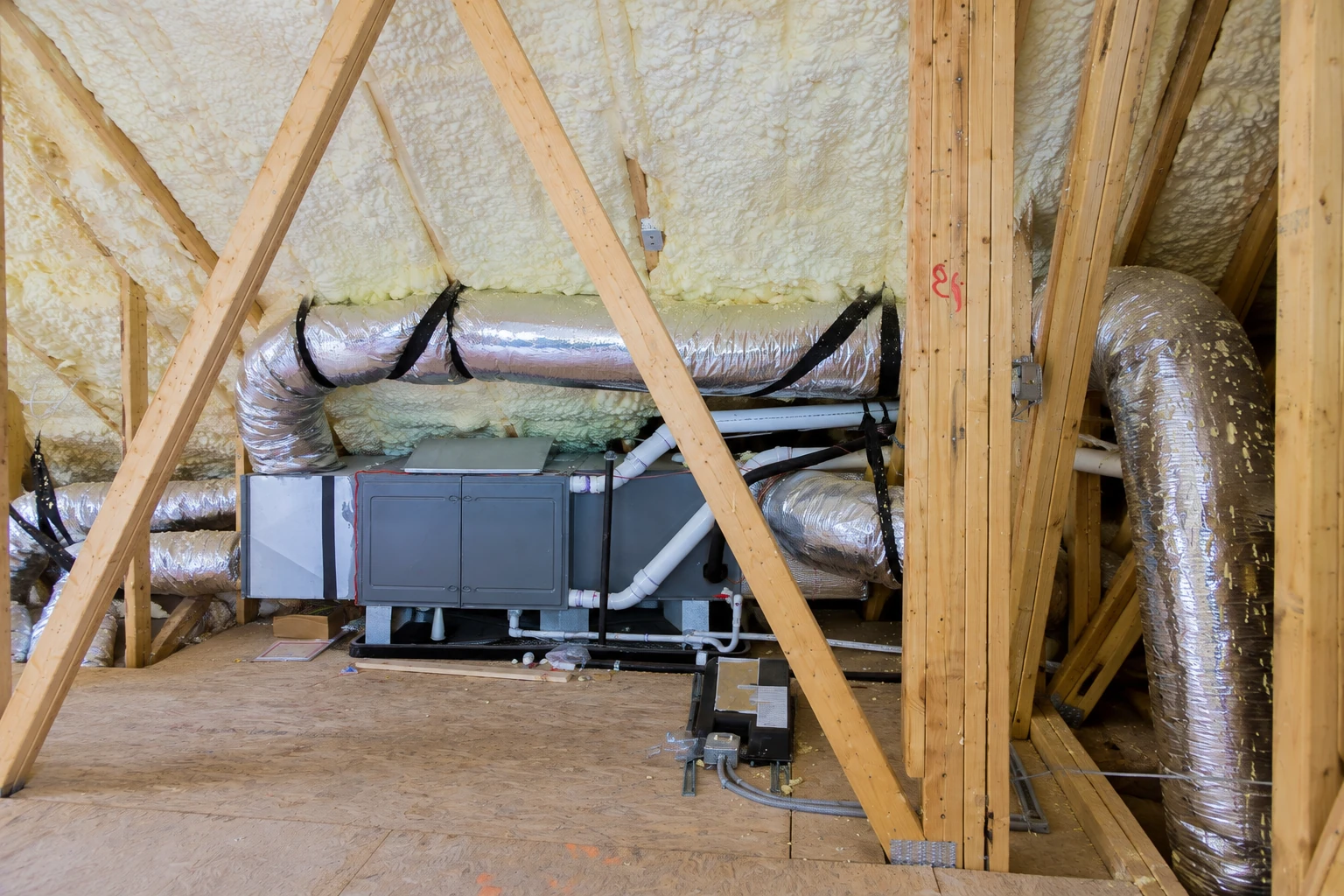 Foam insulation covers the roof and rafters of an attic with a central HVAC unit and silver ducts.