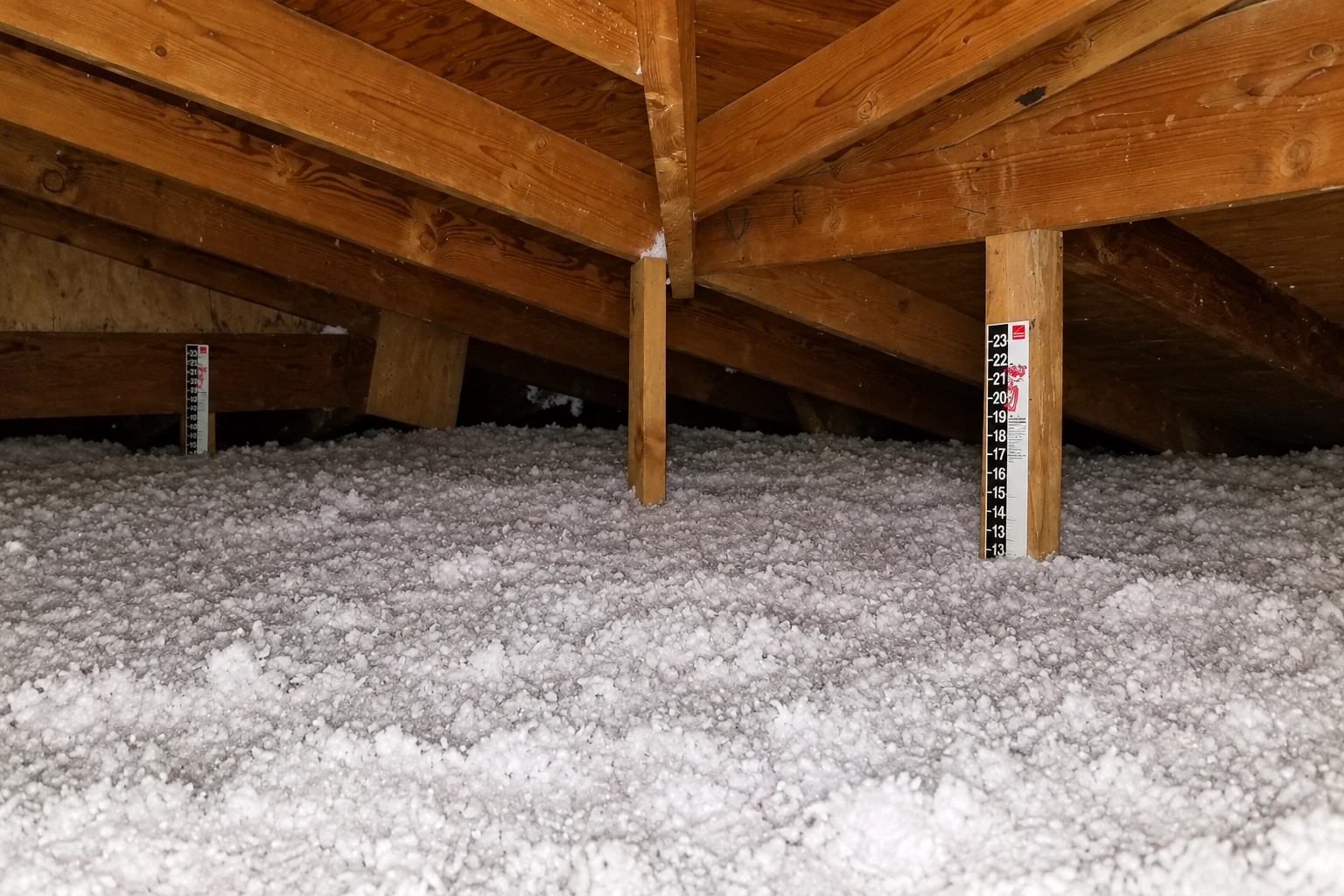 Attic floor covered in thick loose-fill insulation beneath exposed wooden roof trusses, with depth markers standing among the material.