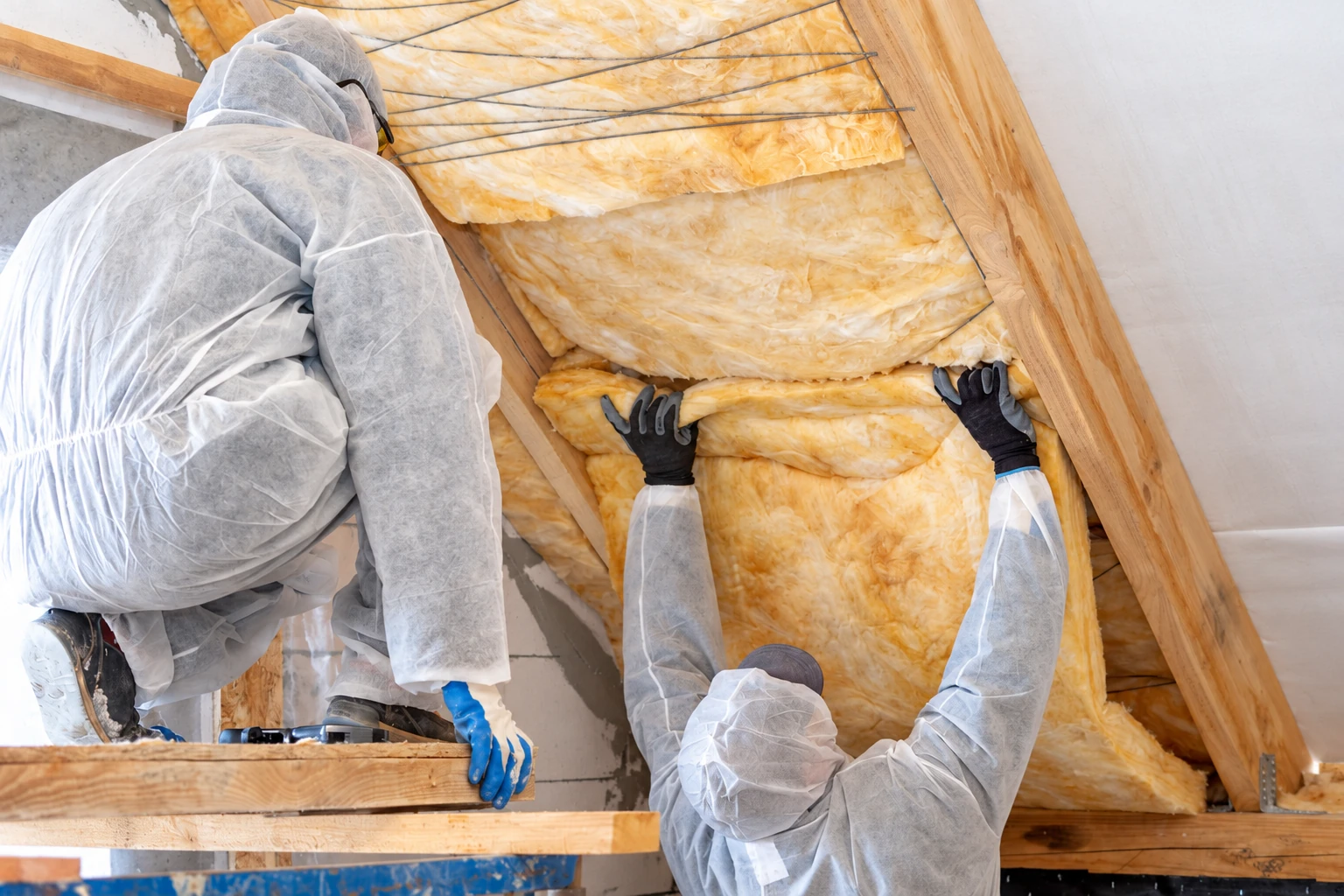 Two workers in protective gear fit yellow fiberglass batts between attic rafters.